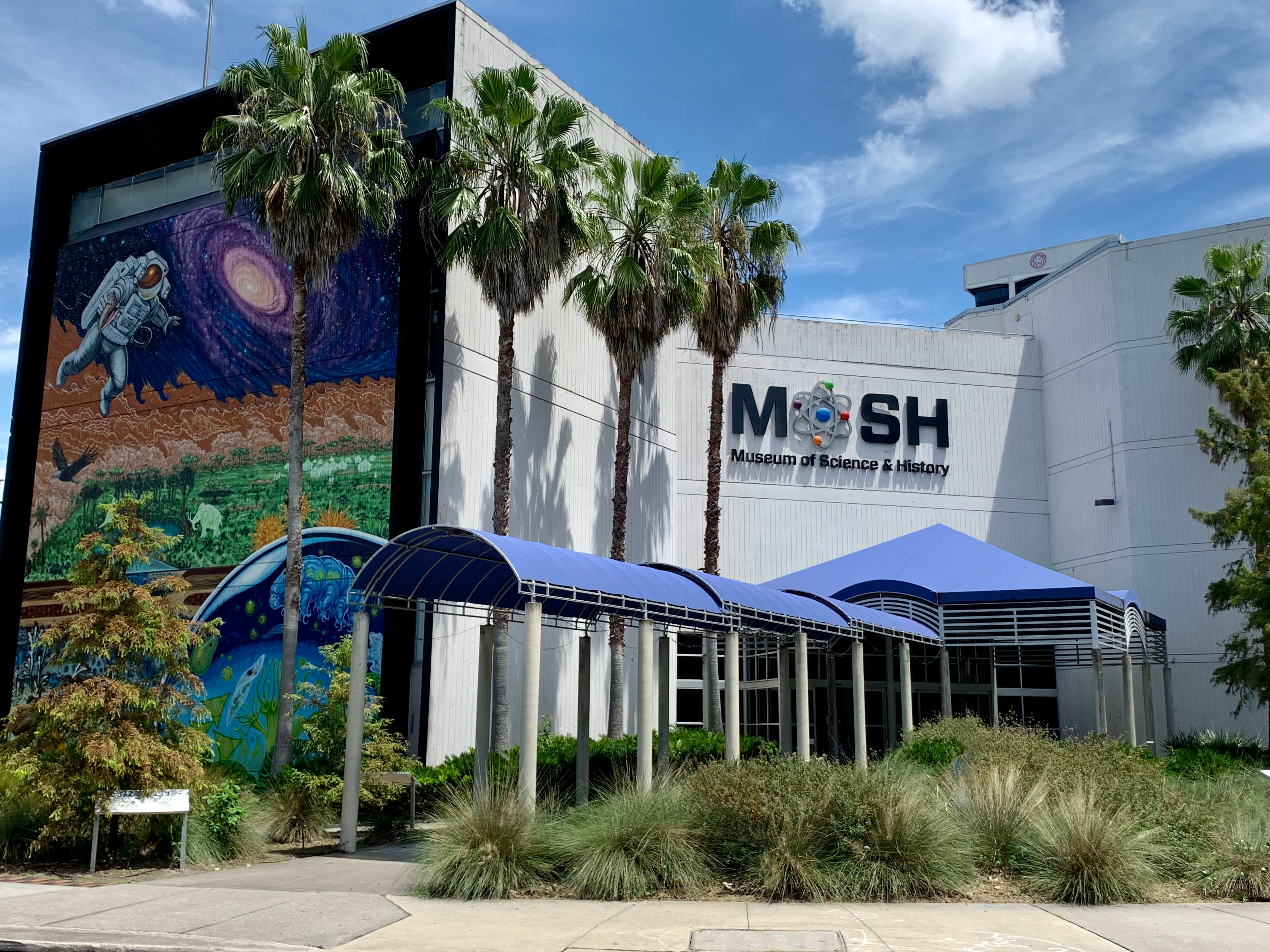 Exterior shot of the MOSH Museum of Science & History. There is a gray building with a covered walkway. To the left of the entrance are palm trees. In front of the entrance is a small dessert garden. The left side of the building has a colorful mural of an astronaut in a space suit floating through space. Below it the mural shows depicts other ecosystems.